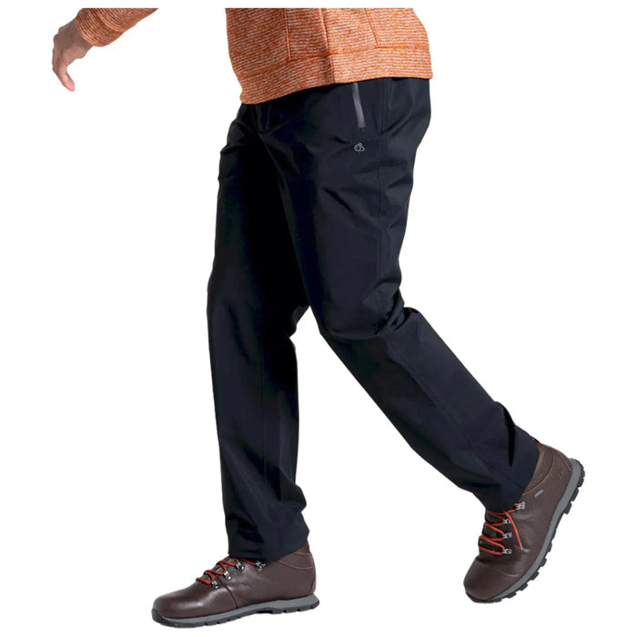 A person is walking with their left leg lifted while wearing black pants and brown hiking shoes. The environment is neutral and uncluttered, focusing on the individual’s movement and attire.