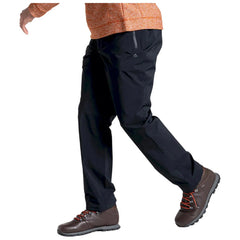 A person is walking with their left leg lifted while wearing black pants and brown hiking shoes. The environment is neutral and uncluttered, focusing on the individual’s movement and attire.